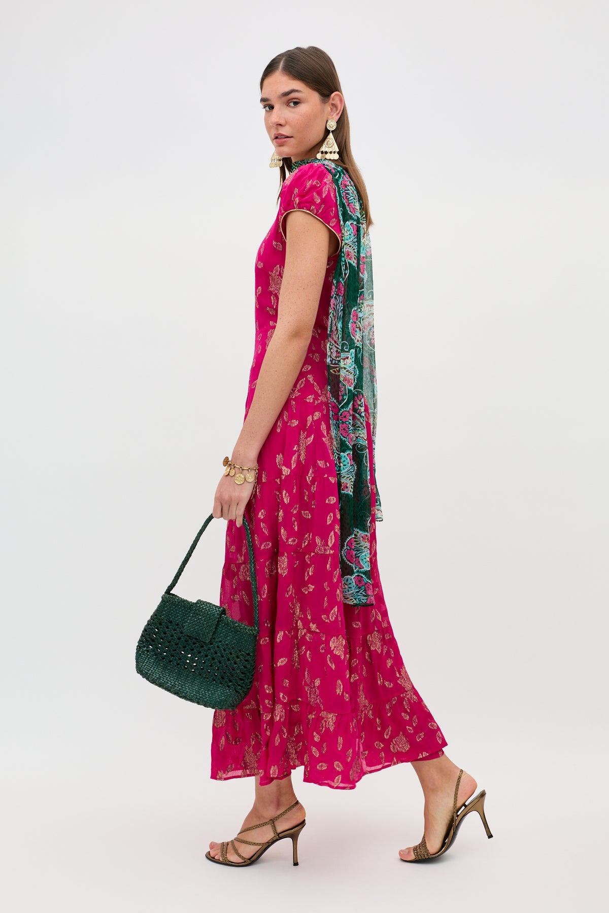A woman wears the Remi - Floral Spark Jacquard Magenta Pink dress with a teal and pink scarf, dark green woven handbag, heeled sandals, gold earrings, and bracelets, posing against a plain white background.