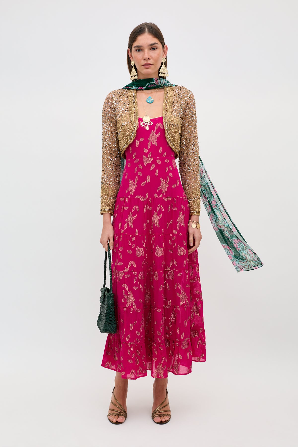 A woman stands against a plain background wearing the Remi - Floral Spark Jacquard Magenta Pink ankle-length dress, a gold embellished cropped jacket, green patterned scarf, dangling earrings, tan sandals, and holding a black purse.