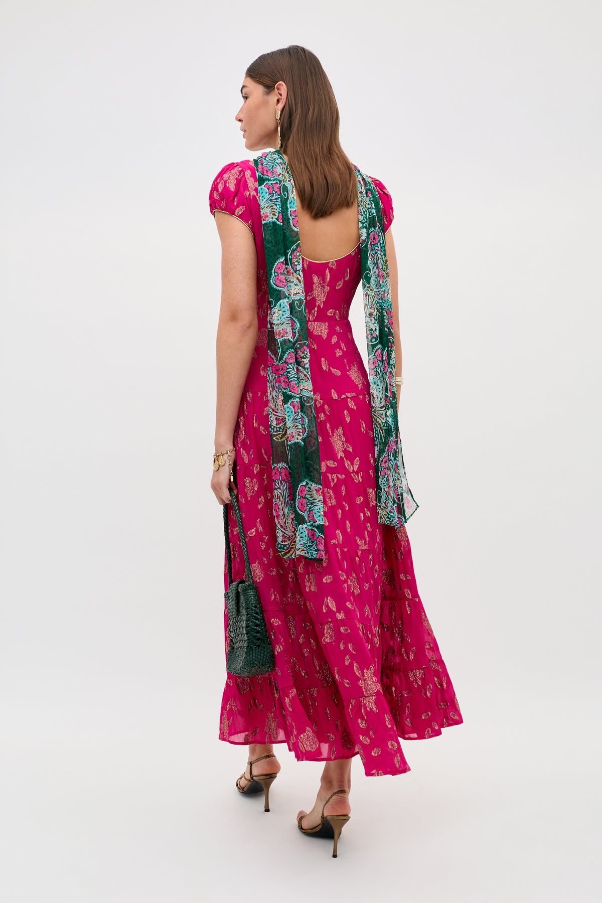 A woman faces away, wearing the Remi - Floral Spark Jacquard Magenta Pink short-sleeve dress with a flared skirt, a green patterned scarf on her shoulders, holding a green bag, and black ankle-strap heels.