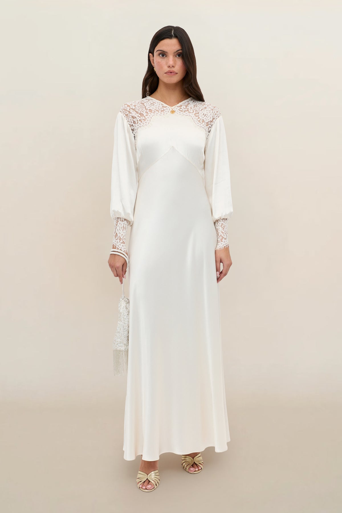 A woman wearing the Axelle – Pearl, a long white satin dress with lace details on the shoulders and sleeves, holds a small purse while standing against a neutral background.