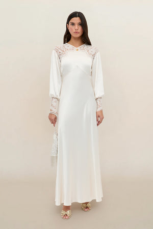 A woman wearing the Axelle – Pearl, a long white satin dress with lace details on the shoulders and sleeves, holds a small purse while standing against a neutral background.
