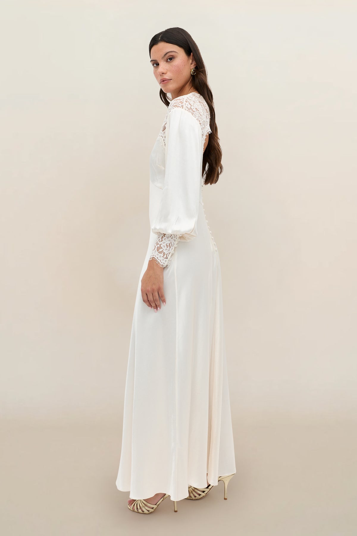 A woman wearing the Axelle - Pearl, a long-sleeved ivory satin gown with lace details, stands sideways and looks at the camera. She has long dark hair, wears light-colored heels, and is set against a plain neutral background.