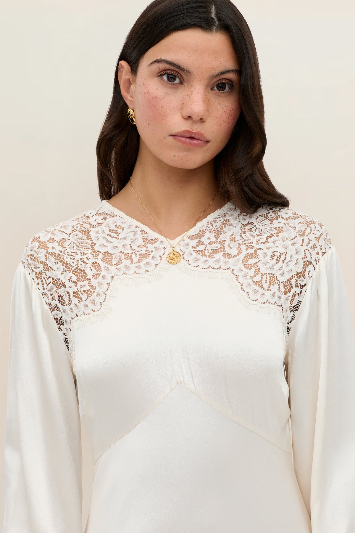 Wearing the Axelle - Pearl ivory dress adorned with lace details on the shoulders and chest, a woman with long brown hair stands against a neutral background, accessorized with gold earrings and a gold necklace.