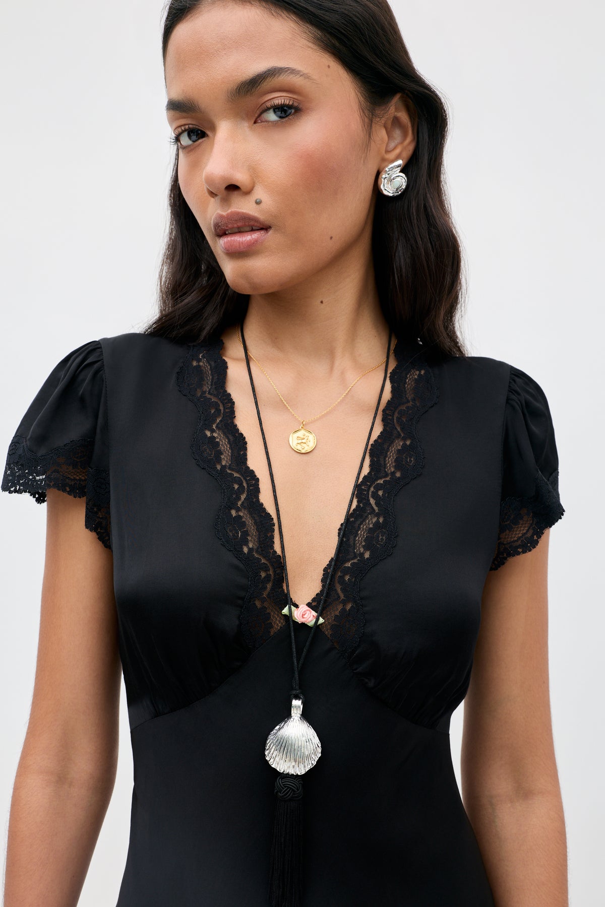 A woman wearing the Cely - Black dress, styled with shell-shaped earrings, a gold necklace, and a long necklace featuring a large silver shell pendant, stands against a plain background.