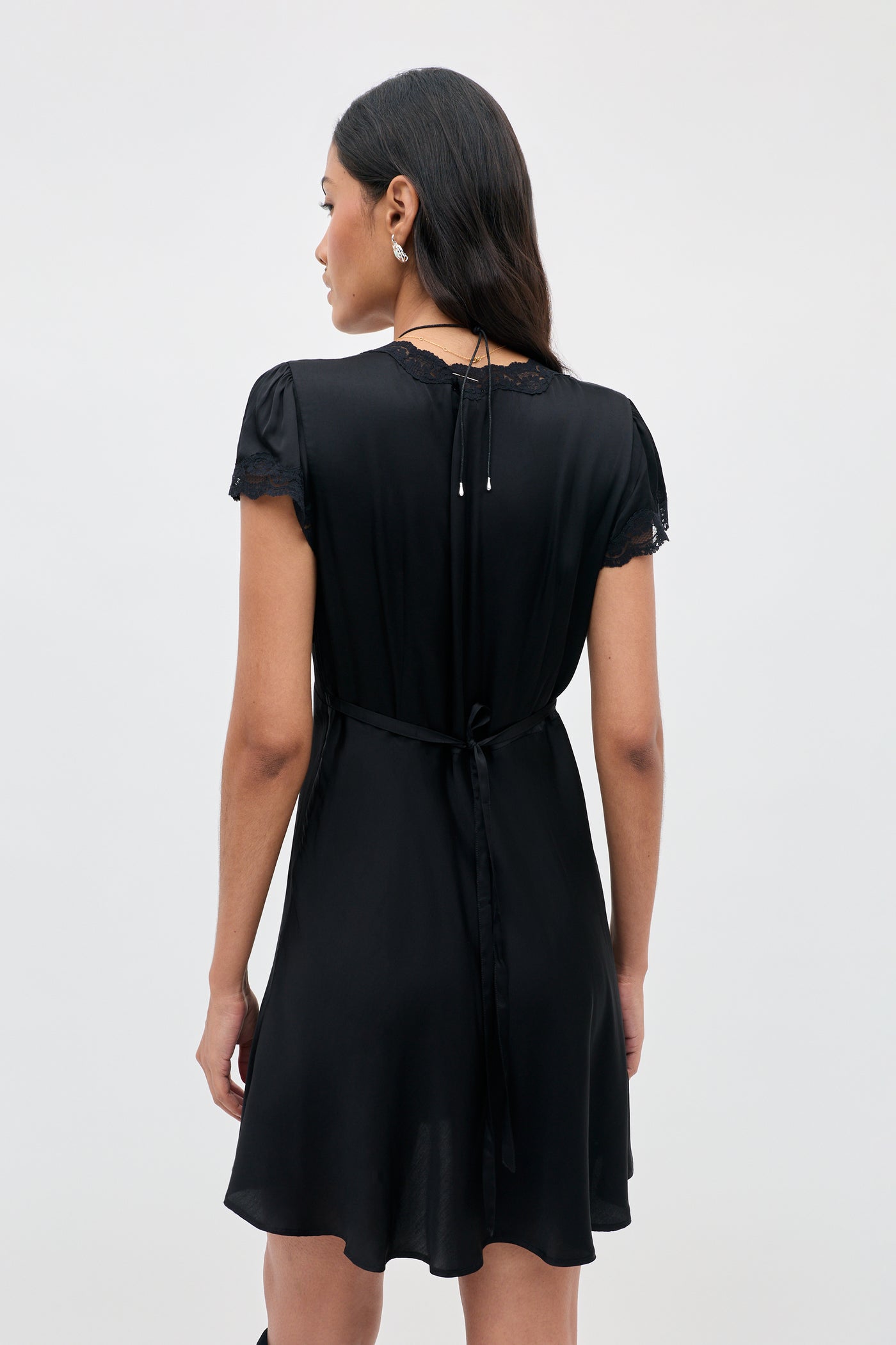 A woman with long dark hair wears the Cely - Black dress, a short black style featuring lace sleeves. She stands facing away to highlight the dress’s back tie detail and its soft, flowing fabric against a plain light background.