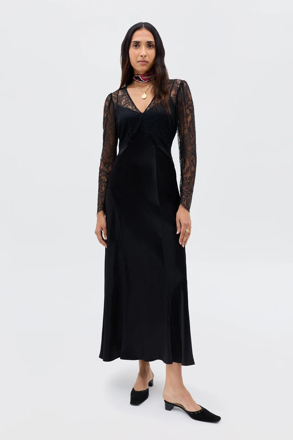 A woman with long dark hair wears the Laoise - Black dress with lace sleeves and black slip-on shoes, posing against a plain white background. She accessorizes with layered necklaces and rings.
