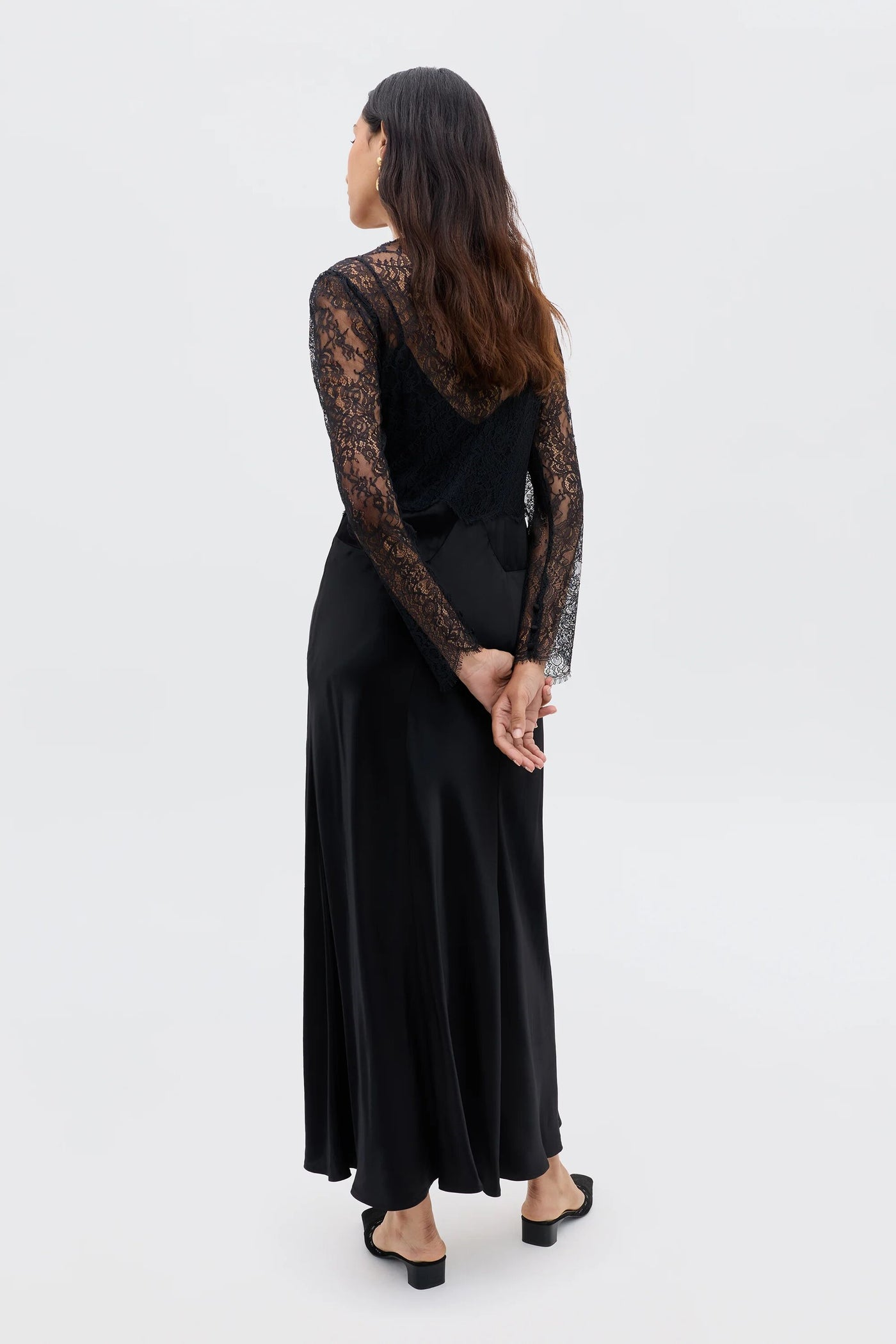 A woman with long brown hair, wearing the Laoise - Black vintage-inspired dress with black lace sleeves and black shoes, stands facing away with her hands clasped behind her back against a plain light background.