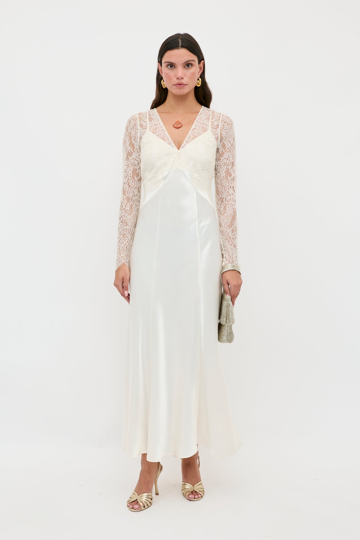 A woman with straight brown hair and a neutral expression stands against a white background wearing the Laoise - Pearl long white satin slip dress with lace sleeves, gold heels, hoop earrings, and holding a small silver clutch.