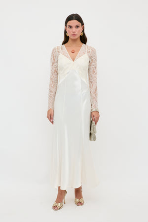 A woman with straight brown hair and a neutral expression stands against a white background wearing the Laoise - Pearl long white satin slip dress with lace sleeves, gold heels, hoop earrings, and holding a small silver clutch.