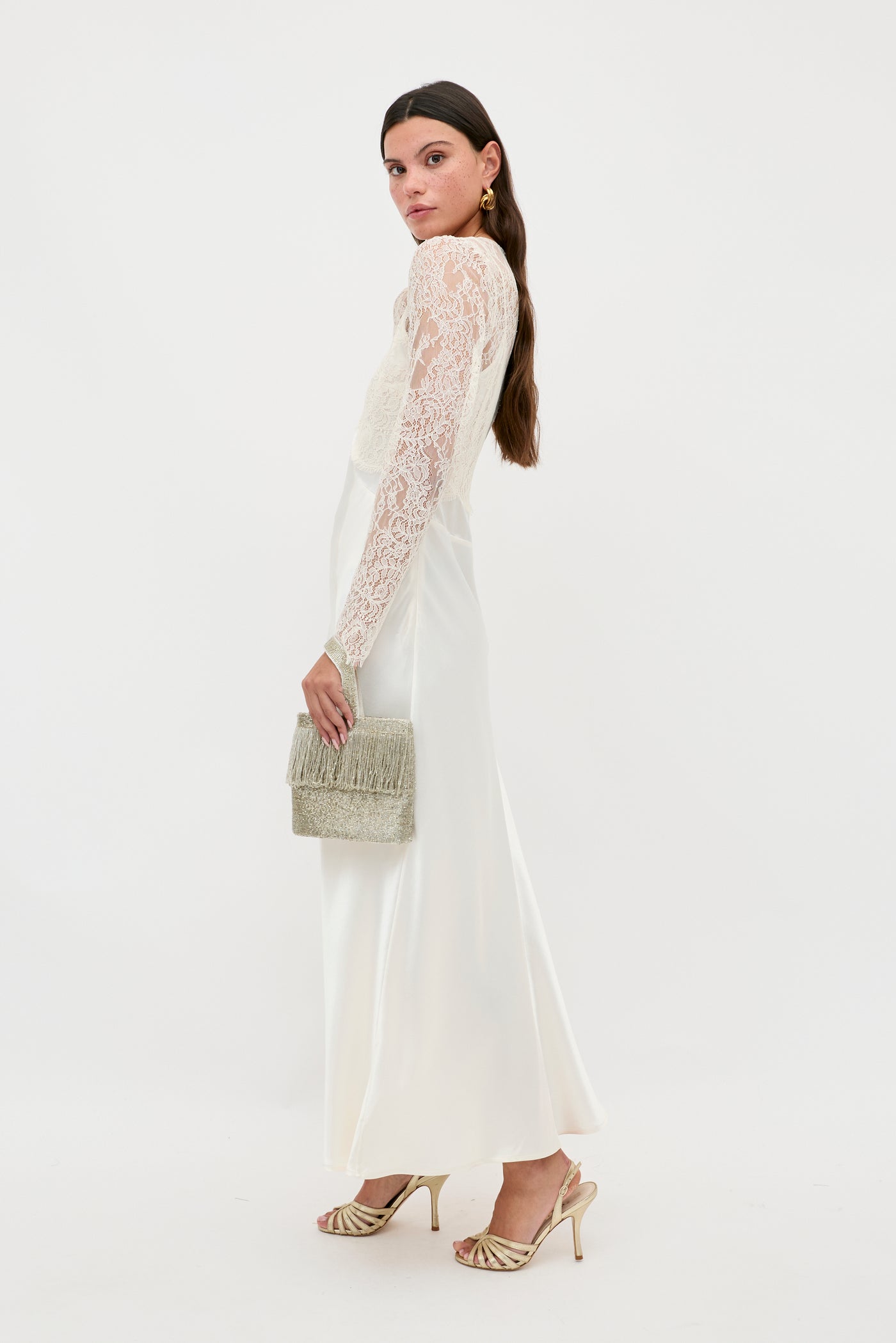 A woman in the Laoise - Pearl long white dress with lace sleeves stands in profile, holding a textured clutch. She wears strappy sandals and gold earrings, her long hair parted in the middle, set against a plain white background.