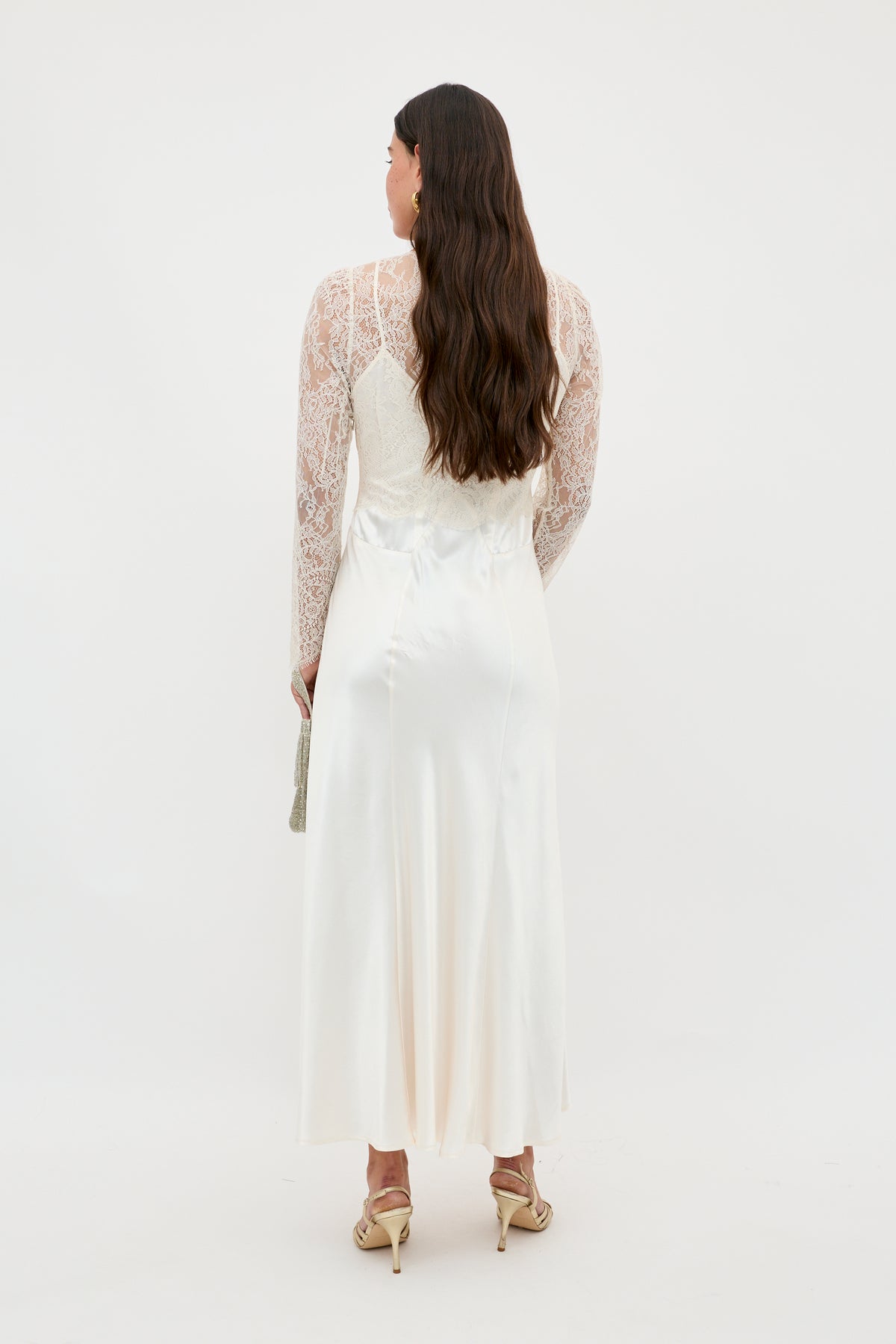 A woman with long wavy brown hair and a small clutch models the Laoise - Pearl, a long white satin dress with lace long sleeves and high heels, standing against a plain white background.