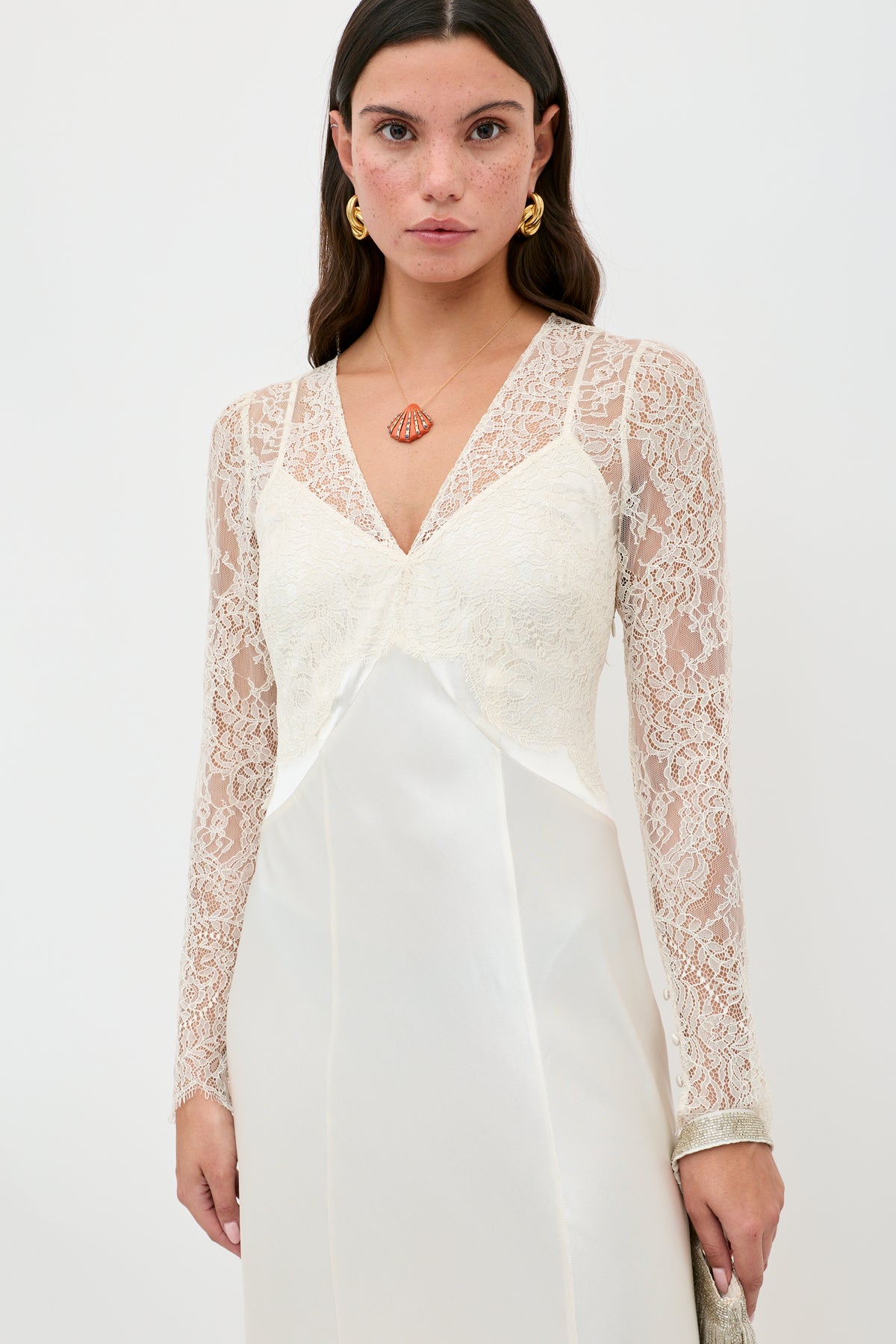 A woman models the Laoise - Pearl dress, a long-sleeved white lace gown with a V-neckline and satin bodice. She has long brown hair and accessorizes with gold earrings and a geometric pendant necklace against a plain background.