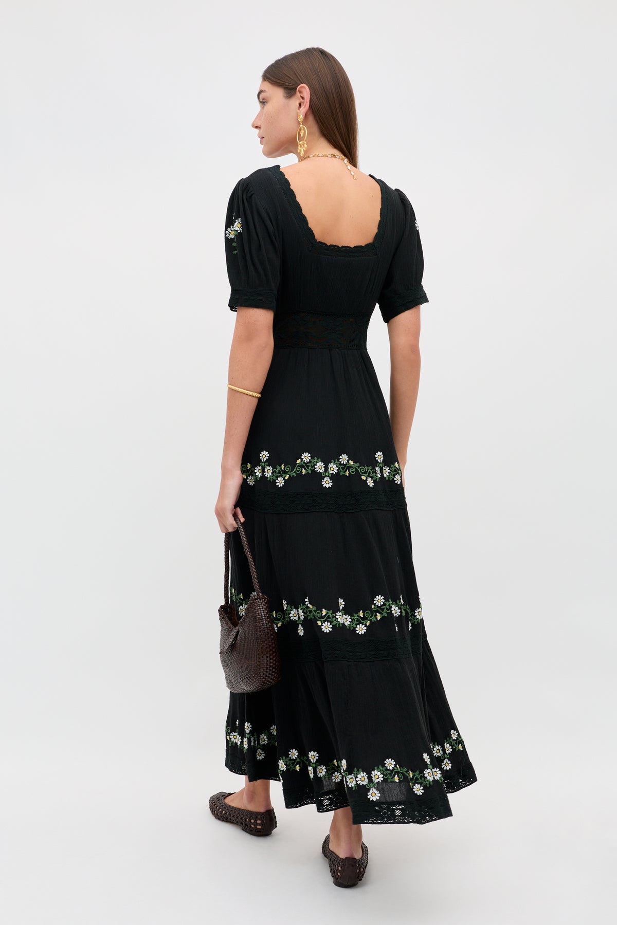 A woman with long brown hair wears the Philippa - Lilia Embroidery Black ankle-length dress, paired with brown woven shoes, gold jewelry, and a brown textured handbag, standing against a plain light background.