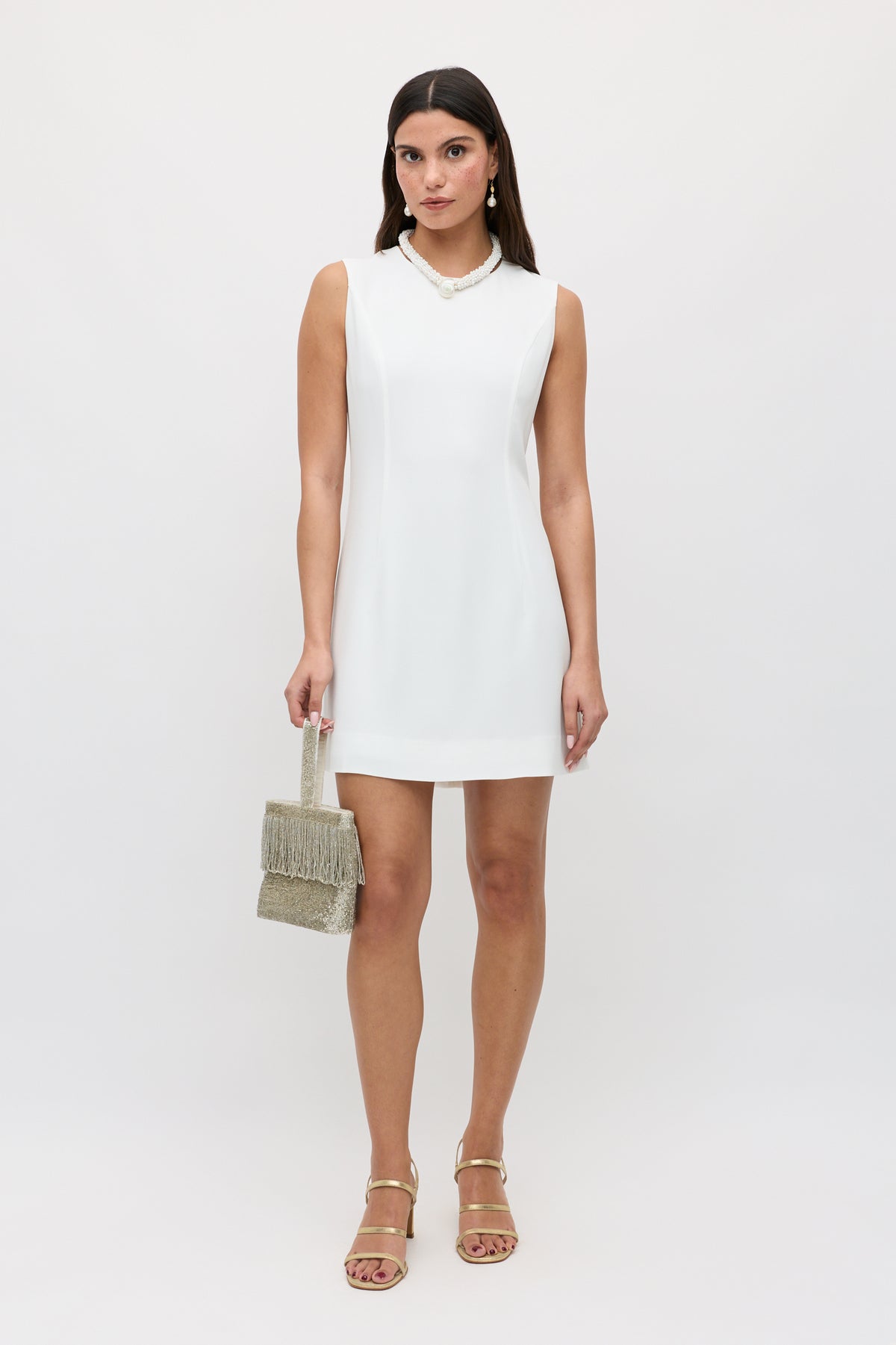 A woman in the sleeveless, boatneck Katrin - Ivory mini dress holds a beaded handbag. She accessorizes with a necklace and gold sandals, and her straight, dark hair contrasts with the light, plain background.