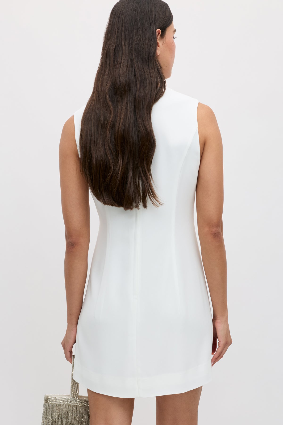 A woman with long brown hair wearing the Katrin - Ivory sleeveless white mini dress with a boatneck stands facing away, holding a textured beige handbag in her left hand against a plain, light background.