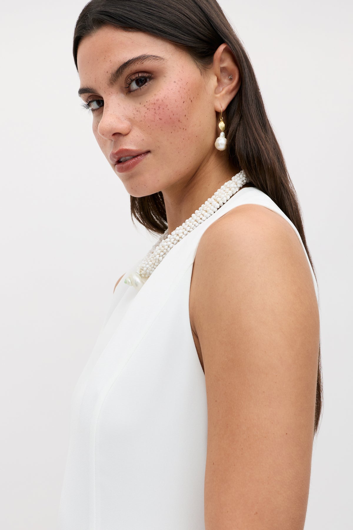 A woman with long dark hair, freckled cheeks, and pearl earrings wears the Katrin - Ivory sleeveless white mini dress featuring a beaded neckline, looking into the camera against a plain light background.