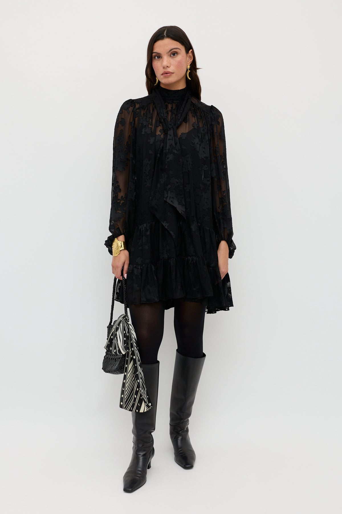 Wearing the Anaya Bouquet Devoré Black dress, a woman stands against a white background paired with black tights, black boots, gold jewelry, a black handbag, and a black-and-white patterned scarf.
