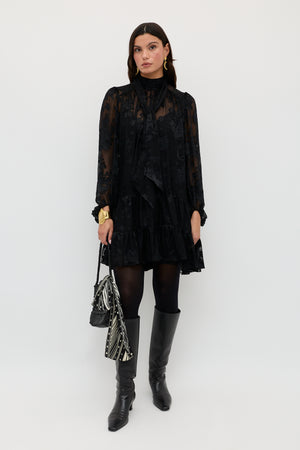 Wearing the Anaya Bouquet Devoré Black dress, a woman stands against a white background paired with black tights, black boots, gold jewelry, a black handbag, and a black-and-white patterned scarf.