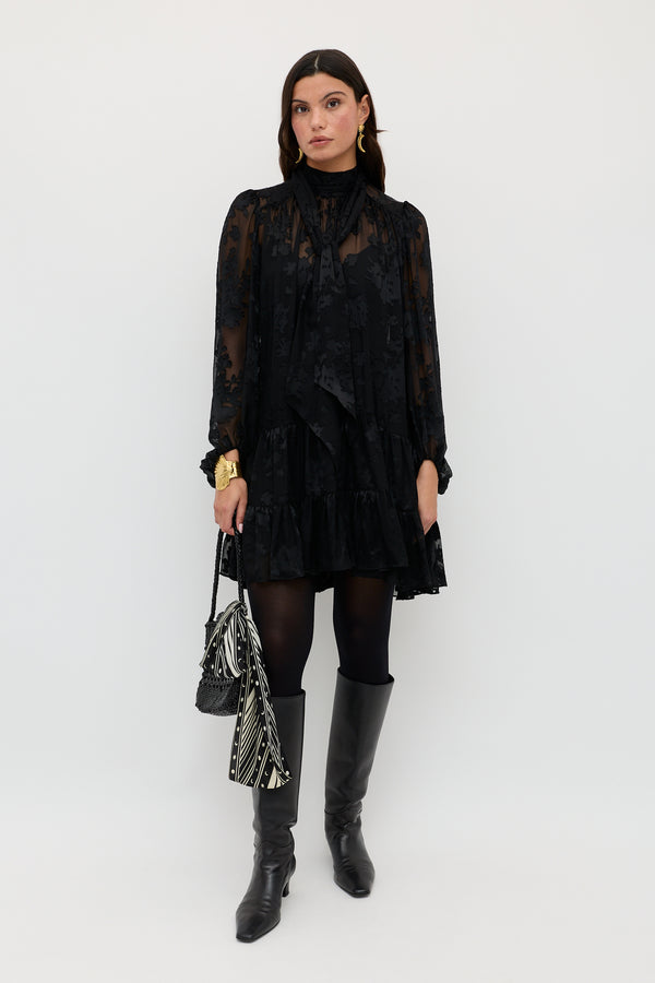 Wearing the Anaya Bouquet Devoré Black dress, a woman stands against a white background paired with black tights, black boots, gold jewelry, a black handbag, and a black-and-white patterned scarf.
