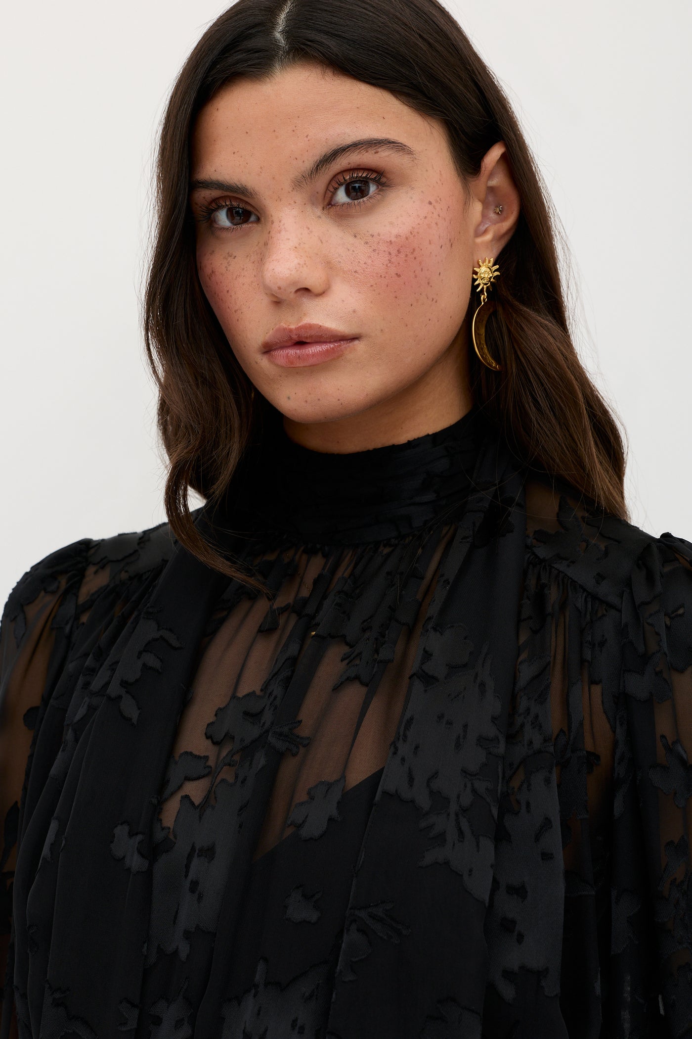 A woman with long brown hair, wearing the Anaya - Bouquet Devoré Black sheer floral-patterned top and gold crescent moon earrings, stands against a plain light background, looking at the camera with a neutral expression.
