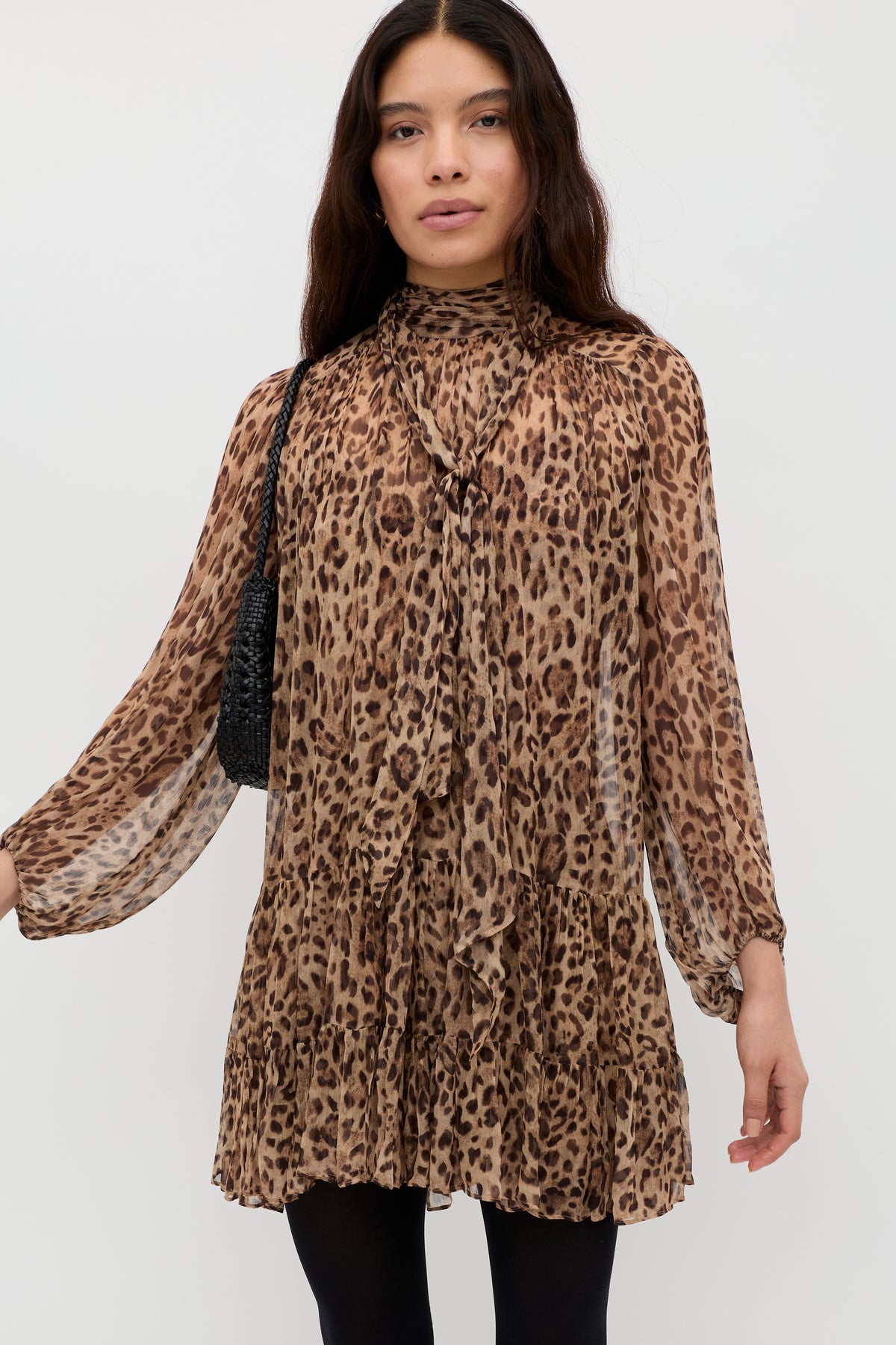 A woman with long dark hair wears the Anaya - Mila Leopard Brown dress, a long-sleeved, above-the-knee leopard print style with a ruffled hem and tied high neckline. She carries a black shoulder bag and stands against a white background.