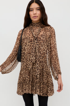 A woman with long dark hair wears the Anaya - Mila Leopard Brown dress, a long-sleeved, above-the-knee leopard print style with a ruffled hem and tied high neckline. She carries a black shoulder bag and stands against a white background.
