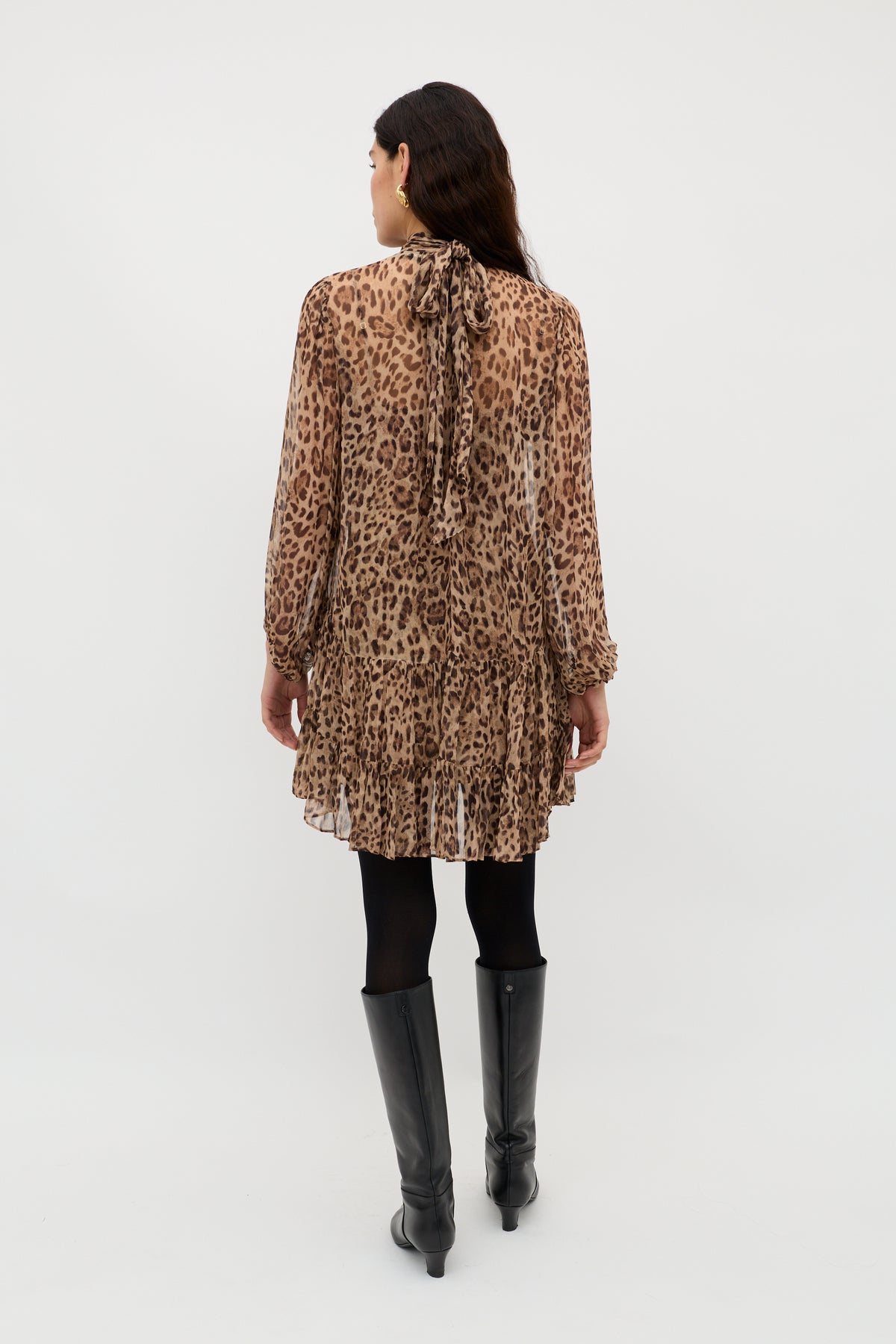 A woman stands facing away, wearing the Anaya - Mila Leopard Brown—a sheer, long-sleeve leopard print dress with a high neckline and ruffled hem, styled with black tights, knee-high boots, and loose dark hair for a bold statement look.