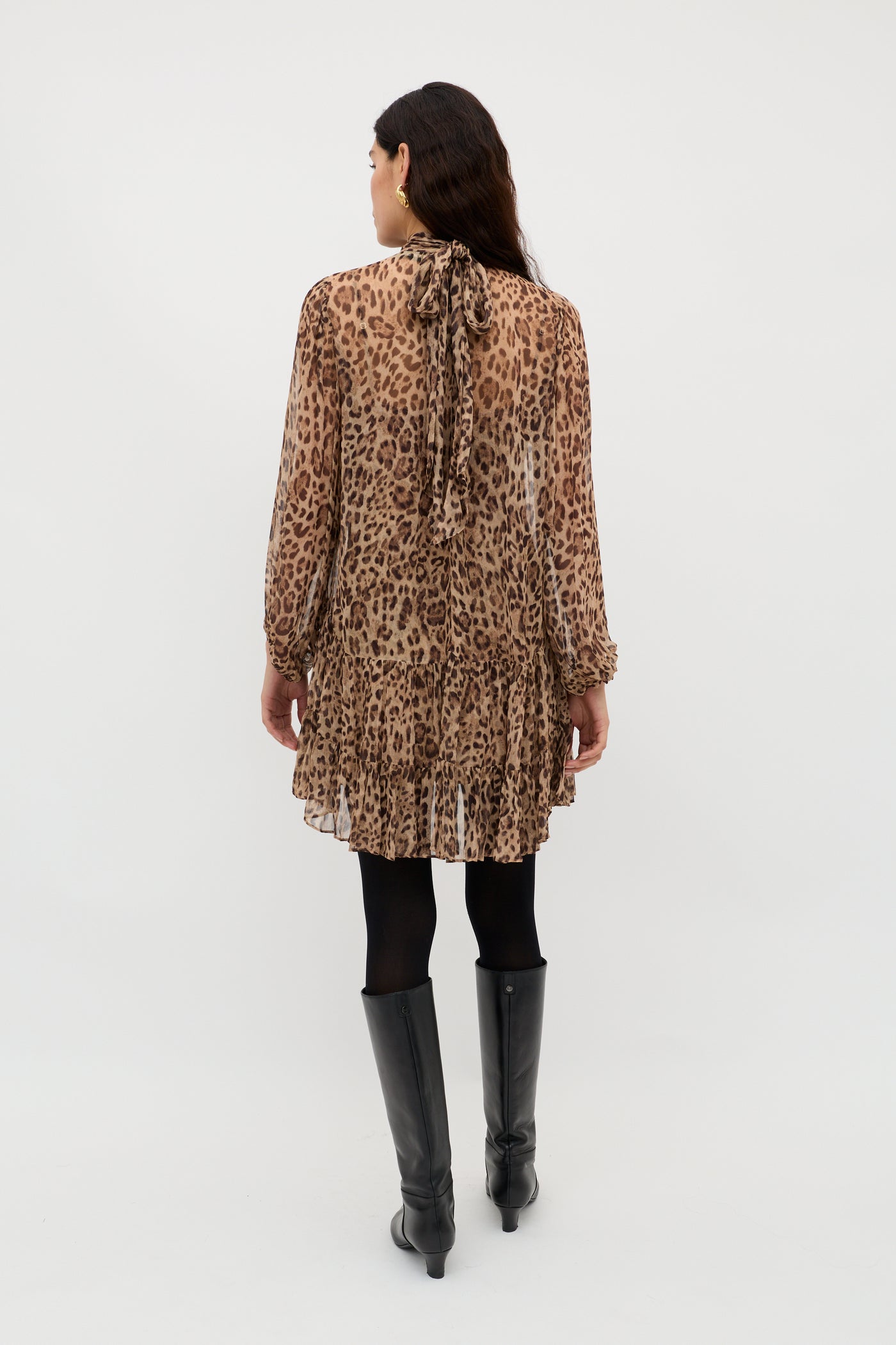 A woman stands facing away, wearing the Anaya - Mila Leopard Brown—a sheer, long-sleeve leopard print dress with a high neckline and ruffled hem, styled with black tights, knee-high boots, and loose dark hair for a bold statement look.