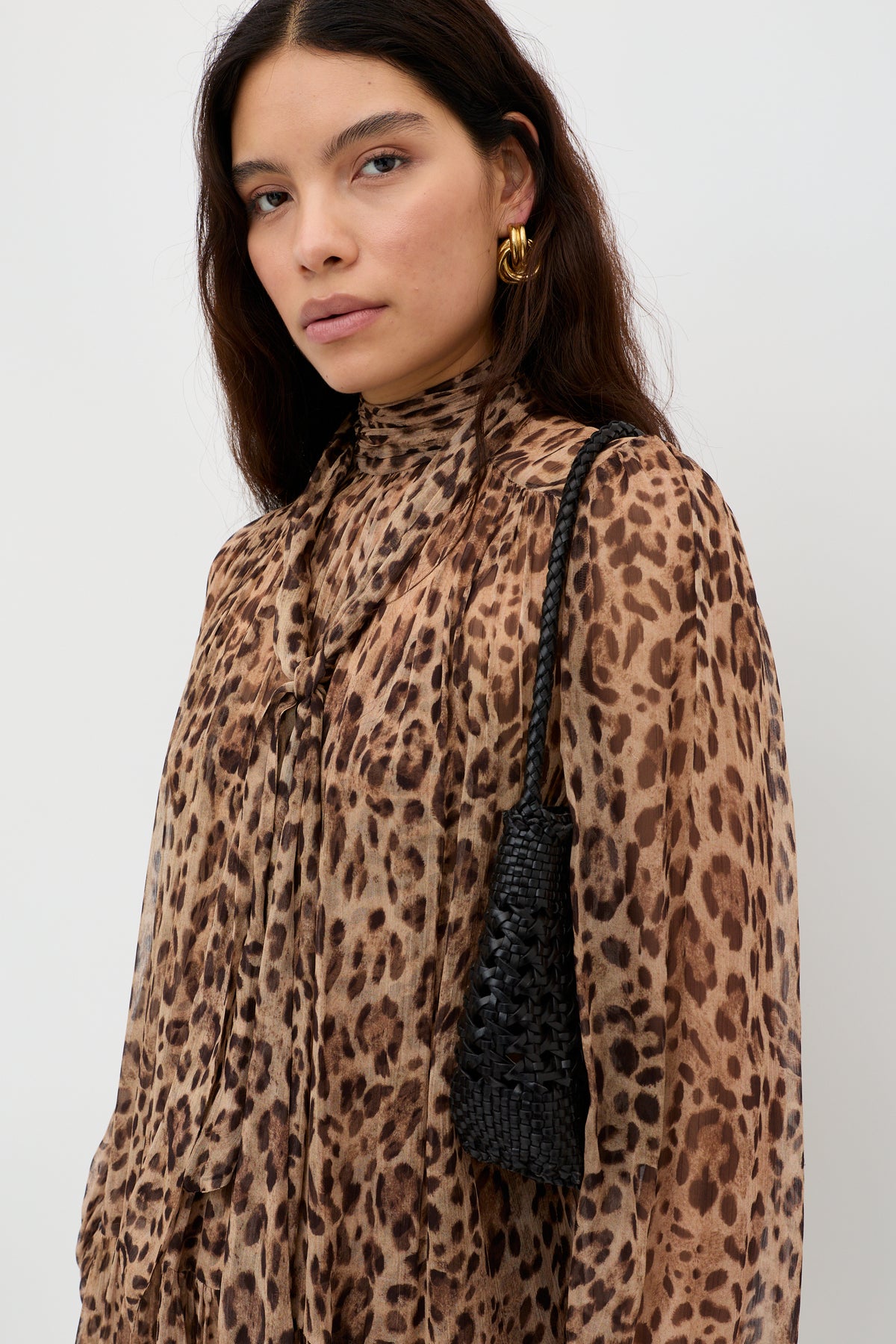 A woman with long dark hair showcases the Anaya - Mila Leopard Brown high-neck, long-sleeve blouse, paired with gold hoop earrings and a small black woven handbag, posing against a plain white background.