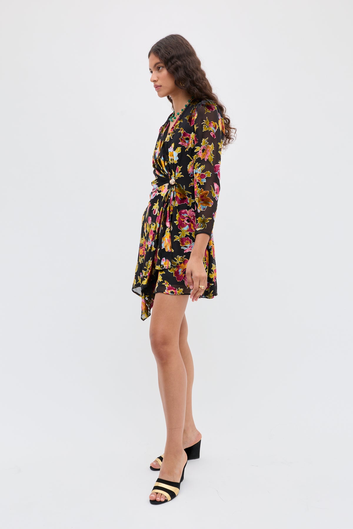 A woman stands sideways in the Kensley - Countryside Devoré Black wrap dress, featuring a colorful print, long sleeves, and a knotted waist, paired with black open-toe low-heeled sandals against a plain white background.