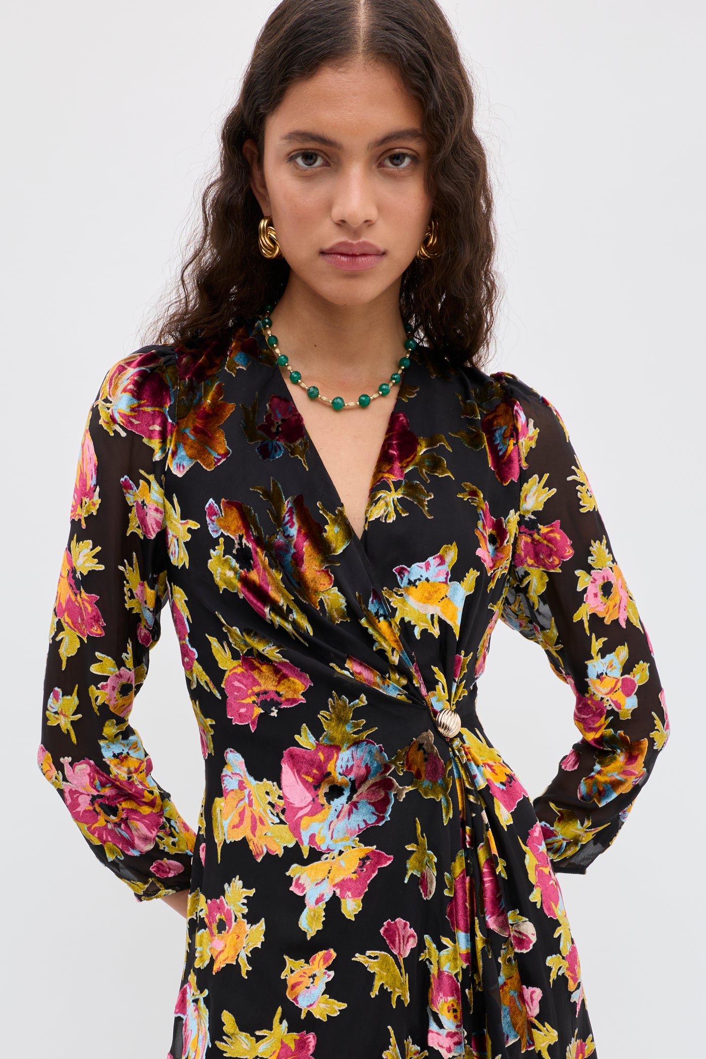 A person with long, wavy hair wears the Kensley - Countryside Devoré Black wrap dress with a floral print, gold hoop earrings, and a green beaded necklace, standing against a plain light background.