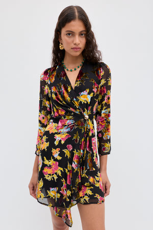 A woman wears the Kensley - Countryside Devoré Black wrap dress with vibrant yellow, pink, and purple floral patterns. She has long curly hair, gold earrings, a green necklace, and stands against a plain white background.