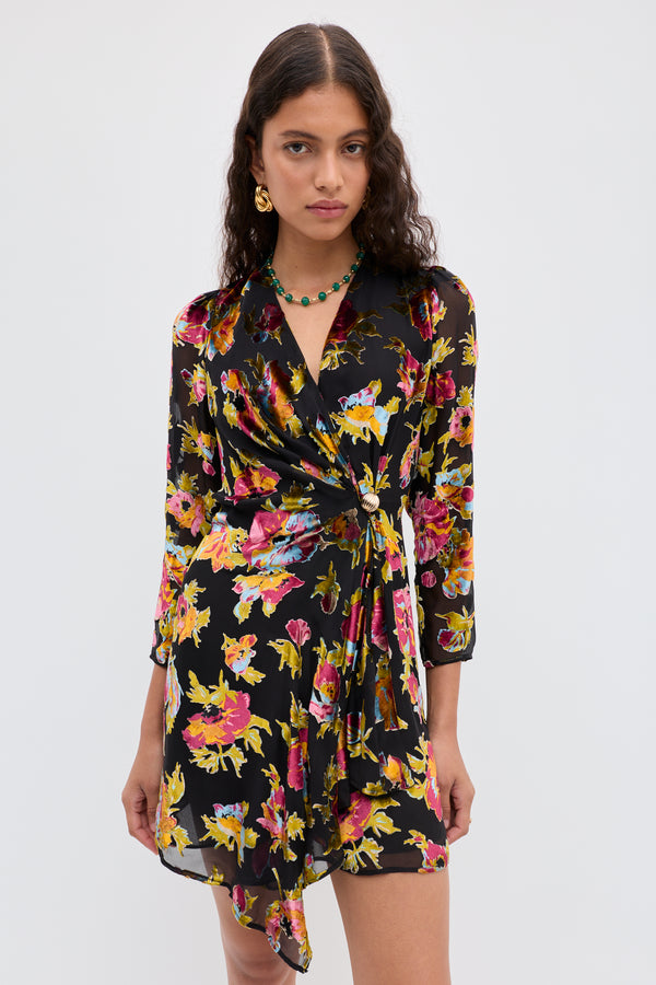 A woman wears the Kensley - Countryside Devoré Black wrap dress with vibrant yellow, pink, and purple floral patterns. She has long curly hair, gold earrings, a green necklace, and stands against a plain white background.