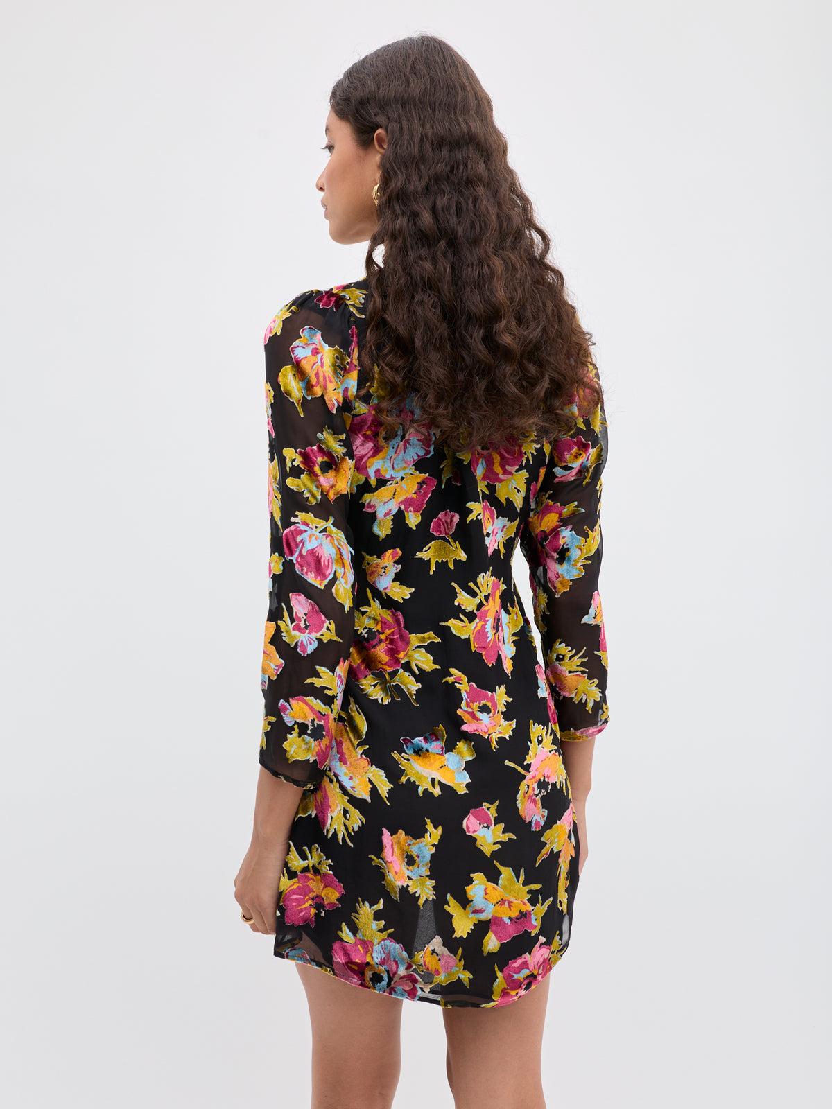 A woman with long, curly brown hair stands with her back to the camera, wearing the Kensley - Countryside Devoré Black dress—a black cocktail dress with colorful floral Devoré, long sleeves, and an above-the-knee hem against a plain white background.