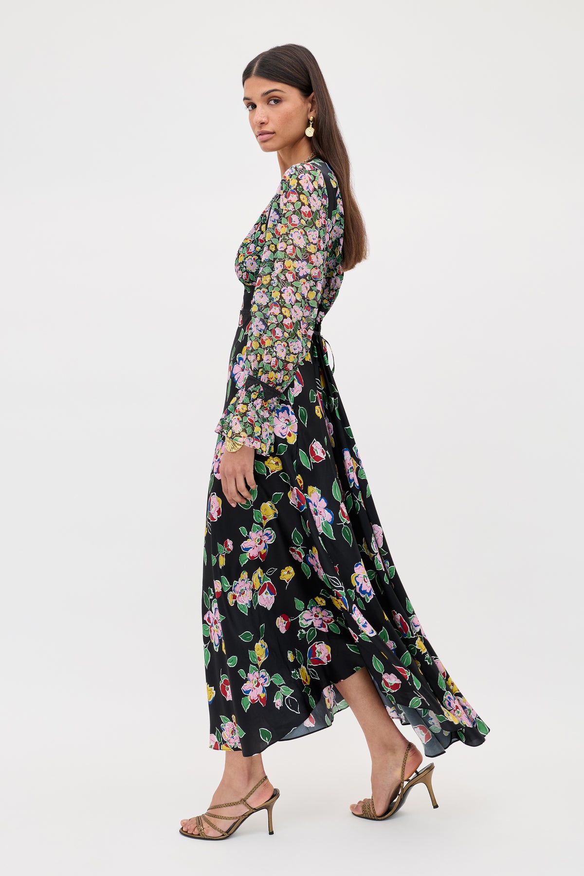 A woman models the Arlena - Kimmie Mix Black dress, a long, flowy black piece with colorful floral prints and sheer floral sleeves. She stands sideways, looking at the camera, paired with strappy sandals and gold earrings.