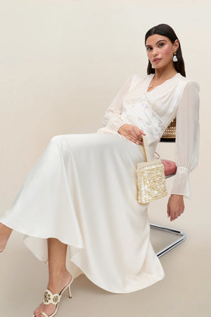 Wearing the Arlena - Pearl long-sleeved satin lace dress, a woman sits on a chair holding a small woven handbag. She pairs the look with white heeled sandals and drop earrings, her dark hair styled straight and sleek against a neutral background.