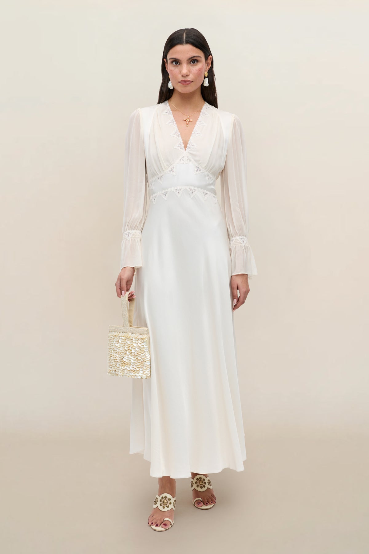 Wearing the Arlena - Pearl dress, a woman stands against a neutral background, showcasing its graceful satin lace design with sheer sleeves. She holds a woven handbag and pairs the look with white sandals. Her long dark hair is styled simply, while earrings and a necklace add an elegant touch—making this ensemble ideal for bridal occasions or special events.