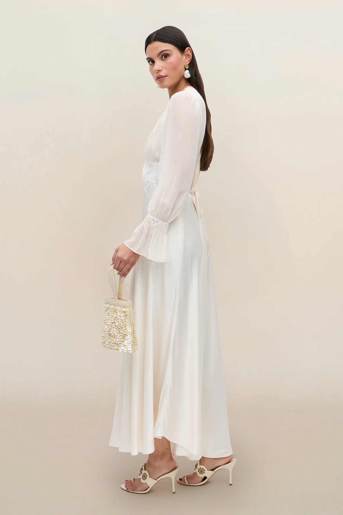 Woman in the Arlena - Pearl long-sleeved, cream-colored wrap dress, holding a small beaded bag and wearing heeled sandals, stands in profile against a light beige background.