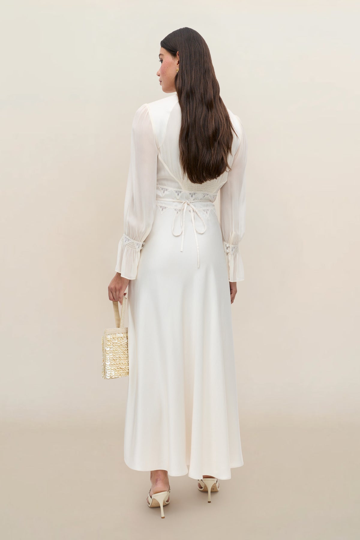 A woman wears the Arlena - Pearl vintage midi dress, featuring long sleeves and a tie at the back, as she stands in heels with a woven handbag, facing away from the camera against a neutral background.