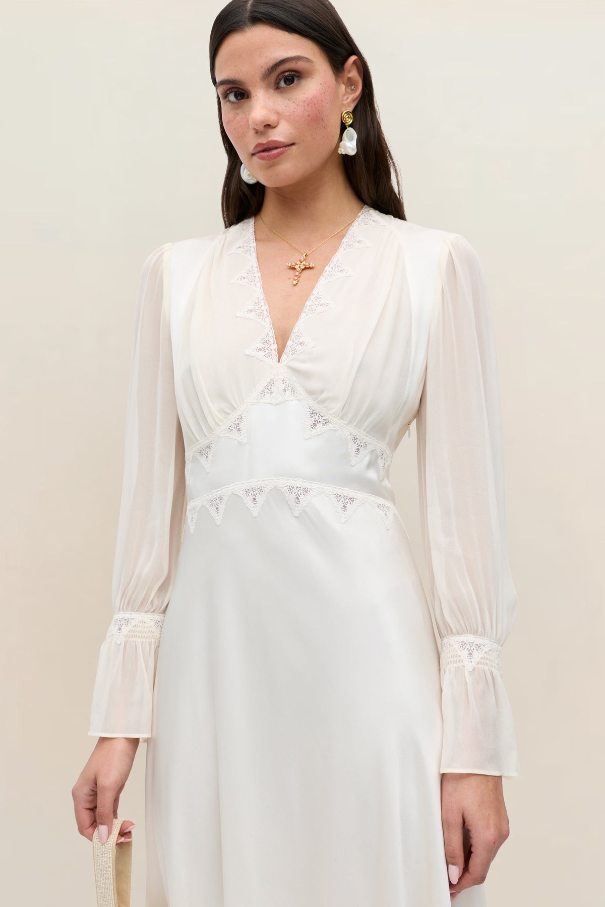 A woman with long brown hair stands against a plain light background, wearing the Arlena - Pearl long-sleeved ivory satin lace dress featuring a V-neckline and delicate lace trim. She accessorizes with a gold necklace, drop earrings, and holds a handbag.