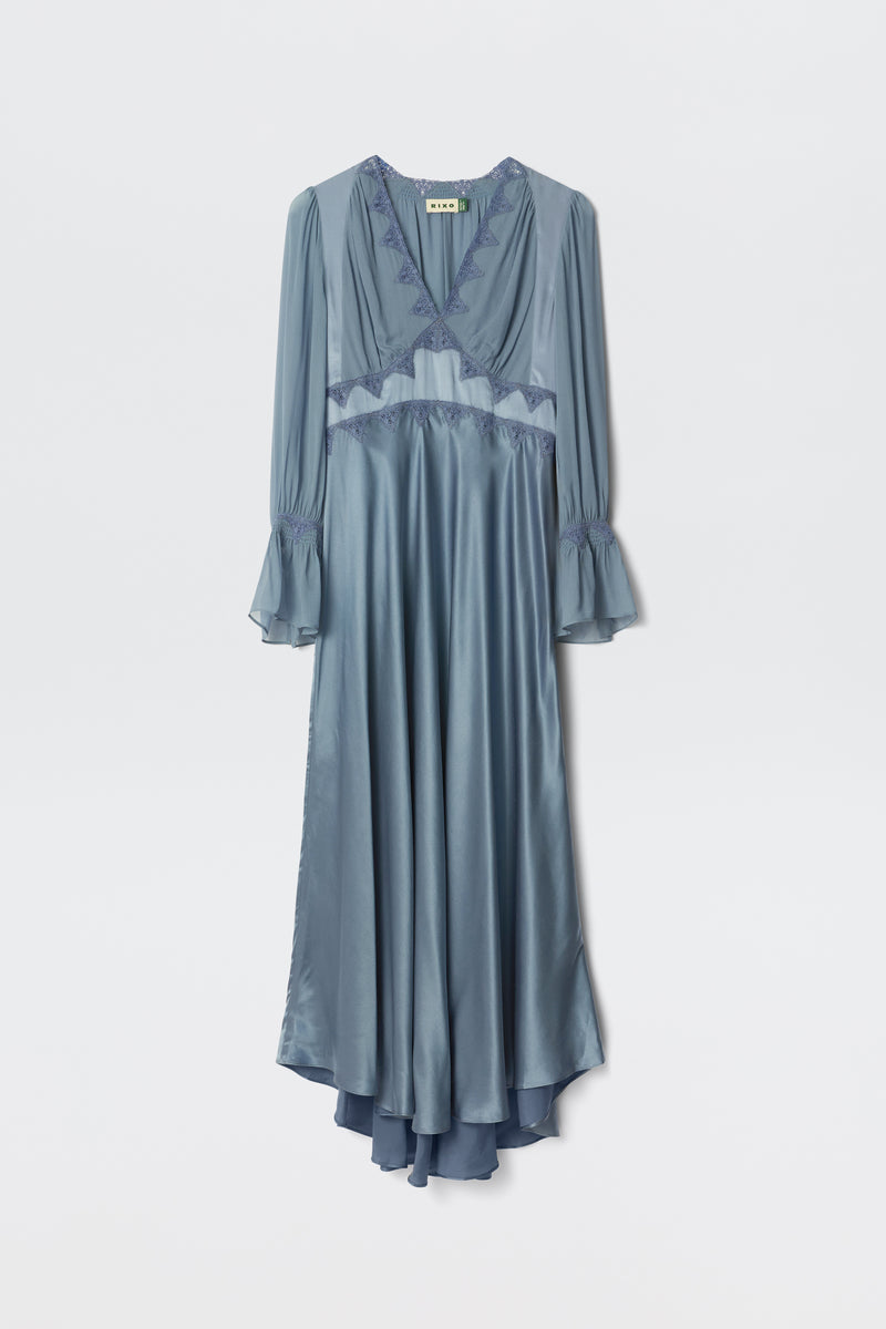 The Arlena in Stone Blue is a long, silky dress with sheer long sleeves, flared cuffs, a V-neckline, lace trim, and an asymmetrical hem, shown against a plain white background.