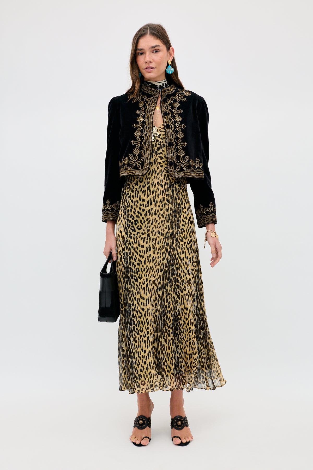 A woman with shoulder-length brown hair wears the Bexley - Nina Leopard Brown dress, a black embroidered jacket, blue earrings, black open-toe sandals, and holds a black handbag. She stands against a plain white background.