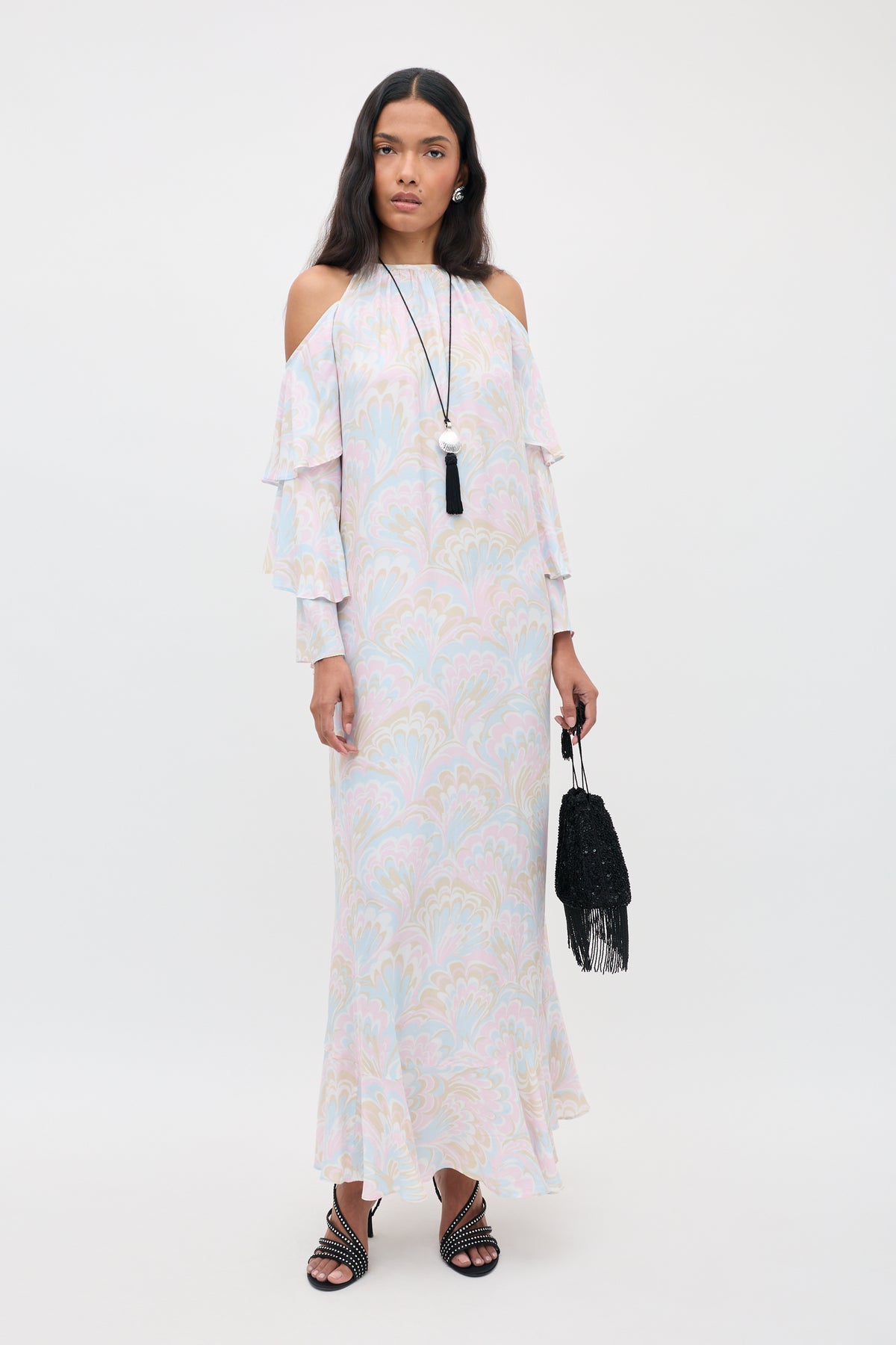 A woman in the Niara - Marble Swirl Pastel cold-shoulder maxi dress with ruffled sleeves, holding a black fringed bag, wearing black sandals and a long necklace, stands against a plain white background.