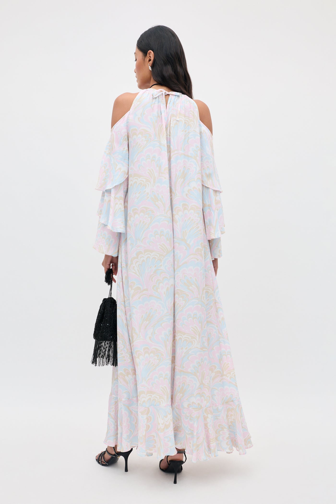 A woman faces away from the camera, wearing the Niara - Marble Swirl Pastel dress with cold shoulder sleeves, paired with a black fringed bag and black heels against a plain background.