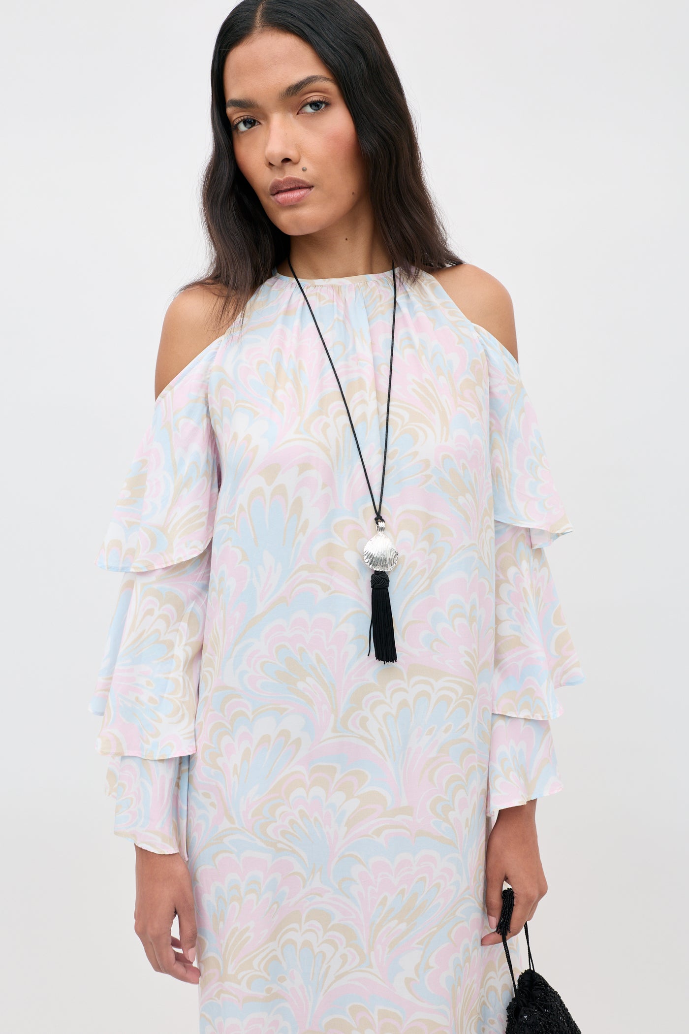 A woman wearing the Niara - Marble Swirl Pastel cold-shoulder dress with ruffled sleeves and a long black necklace stands against a plain light background.