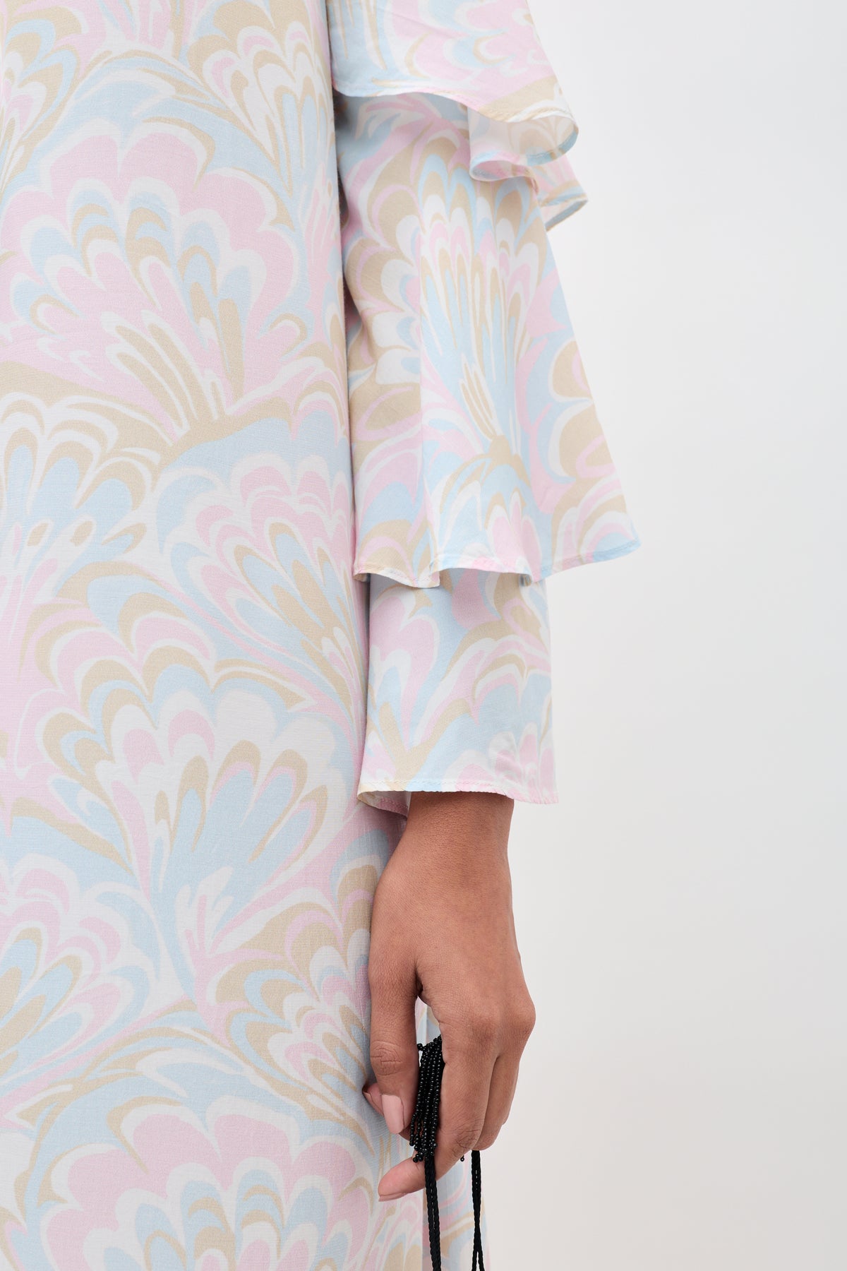A person wearing the Niara - Marble Swirl Pastel dress with ruffled sleeves, holding a black beaded accessory. Only the lower arm and part of the dress are visible against a plain light background.