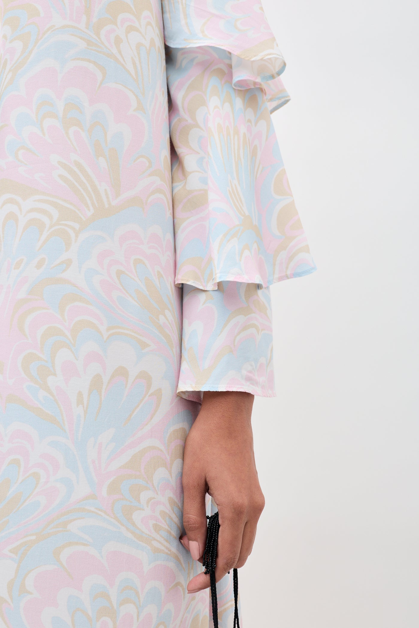 A person wearing the Niara - Marble Swirl Pastel dress with ruffled sleeves, holding a black beaded accessory. Only the lower arm and part of the dress are visible against a plain light background.