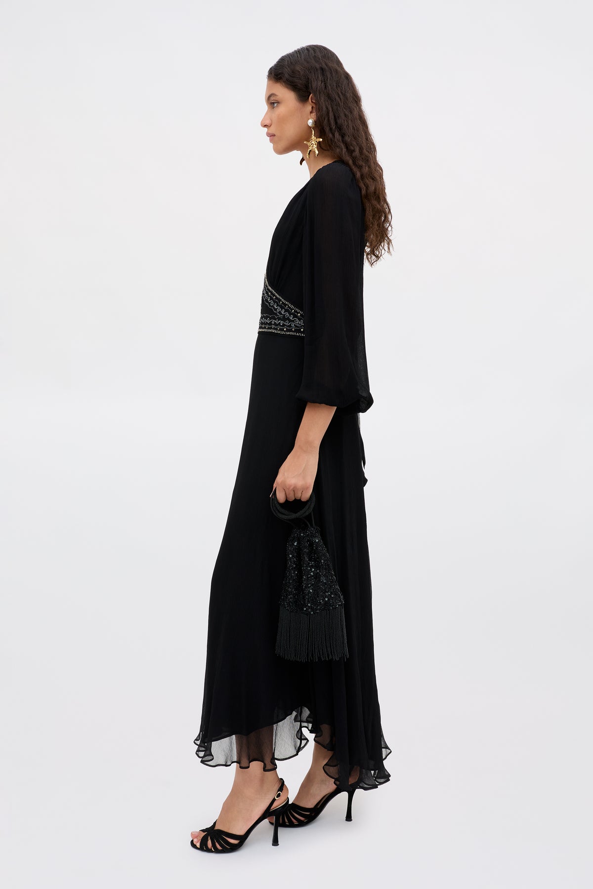 Wearing the Kaesha - Diana Embellishment Black, a woman with long, wavy hair and star-shaped earrings stands against a light background. She carries a black beaded fringe purse and wears black heeled sandals.