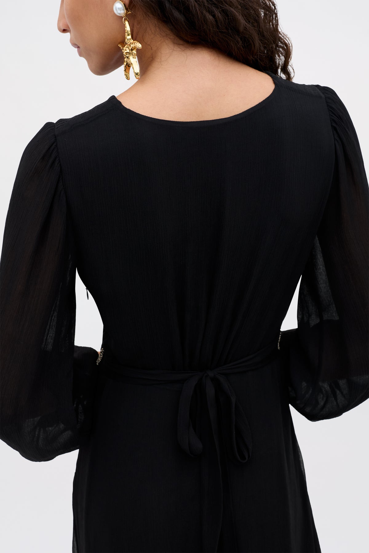 A woman with long, wavy brown hair models the Kaesha - Diana Embellishment Black dress, featuring sheer long sleeves and a tie waist. She accessorizes with large gold statement earrings adorned with pearls.