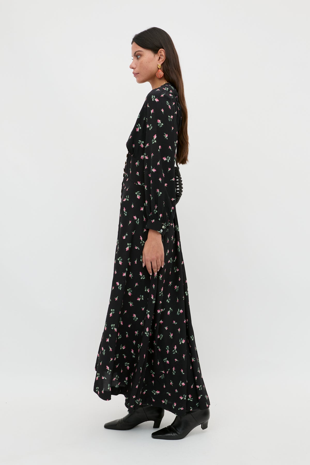 A woman in profile against a white background wears the Avalyn - Sienna Bud Black long sleeve midi dress featuring a floral pattern and vintage-inspired cuffs, paired with black ankle boots. She has long dark hair and earrings.
